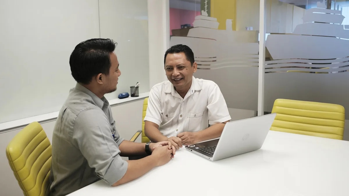 Dua profesional bisnis Indonesia berdiskusi di ruang meeting kantor dengan laptop untuk meninjau keputusan operasional.